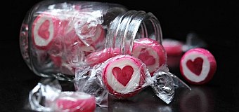 Zdjęcie przedstawia cukierki wysypujące się ze szklanego słoika. Słoik leży na czarnym blacie. Cukierki są białe z dużym, czerwonym sercem pośrodku.