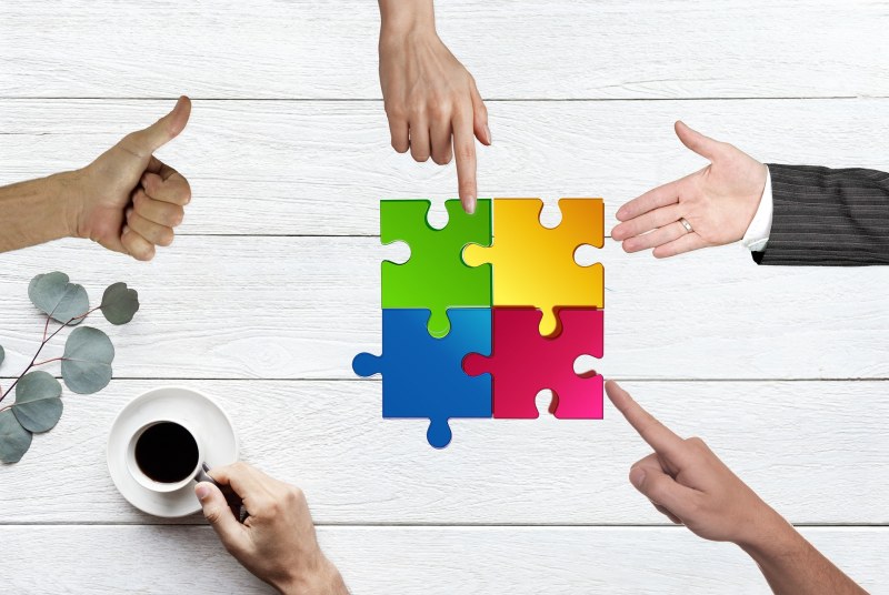 cztery kolorowe puzzle, wokół nich pięć gestykulujących dłoni sugerujących spotkanie biznesowe