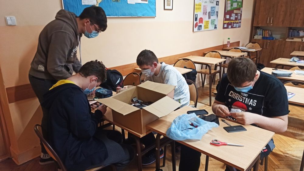 4 chłopcy z klasy 8d i 8e. Uczniowie usuwają baterie z uszkodzonych telefonów komórkowych. Uczniowie są w klasie, siedzą przy stolikach lekcyjnych. Na stoliku stoi pudełko z telefonami, które udało się zebrać podczas akcji Ekophone