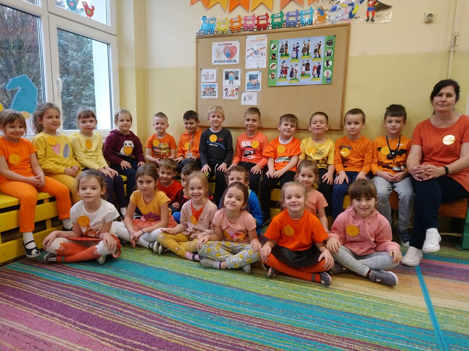 przedstawia grupę 0e, dzieci siedzą z wychowawcą, wszyscy ubrani na pomarańczowo