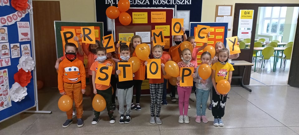 Zdjęcie 6 – przedstawia klasę 1c, dzieci stoją na korytarzu, wszyscy są ubrani na pomarańczowo, w dłoniach trzymają pomarańczowe balony lub pomarańczowe kartki z literami tworzącymi hasło STOP PRZEMOCY