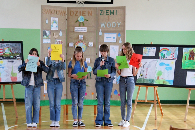 Na zdjęciu uczennice klasy 7. Uczennice ubrane w  niebieskich kolorach , mają na sobie jeansowe spodnie i bluzy oraz niebieskie lub białe koszulki. Przedostatnia dziewczynka od lewej trzyma mikrofon, wszystkie uczennice posiadają w rękach kolorowe kartki.