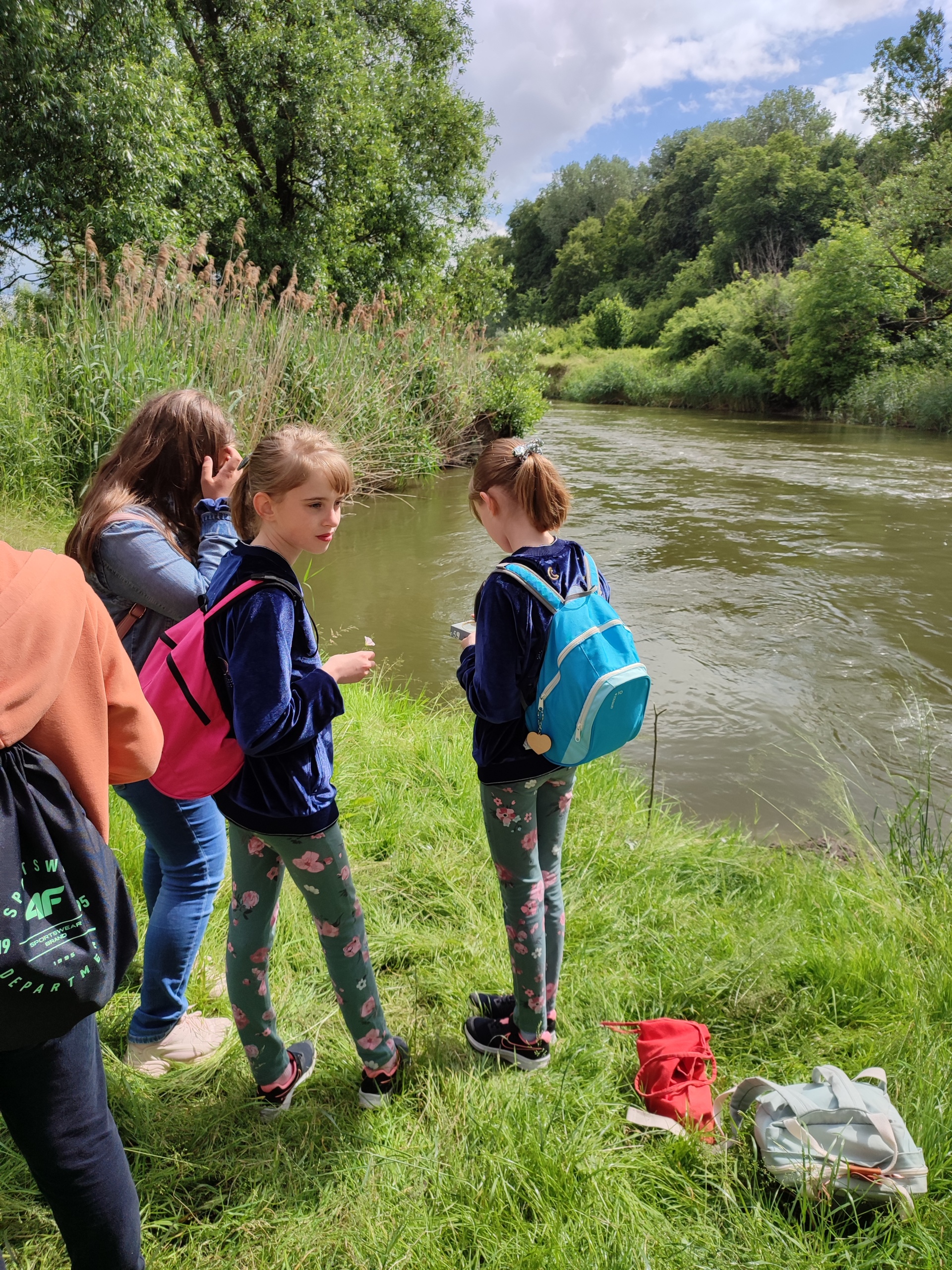 Czterech uczniów w różnokolorowych strojach z plecakami stoi nad brzegiem rzeki.W oddali widać zielone drzewa, krzewy, w pobliżu rzeki zielona trawę i sitowie.