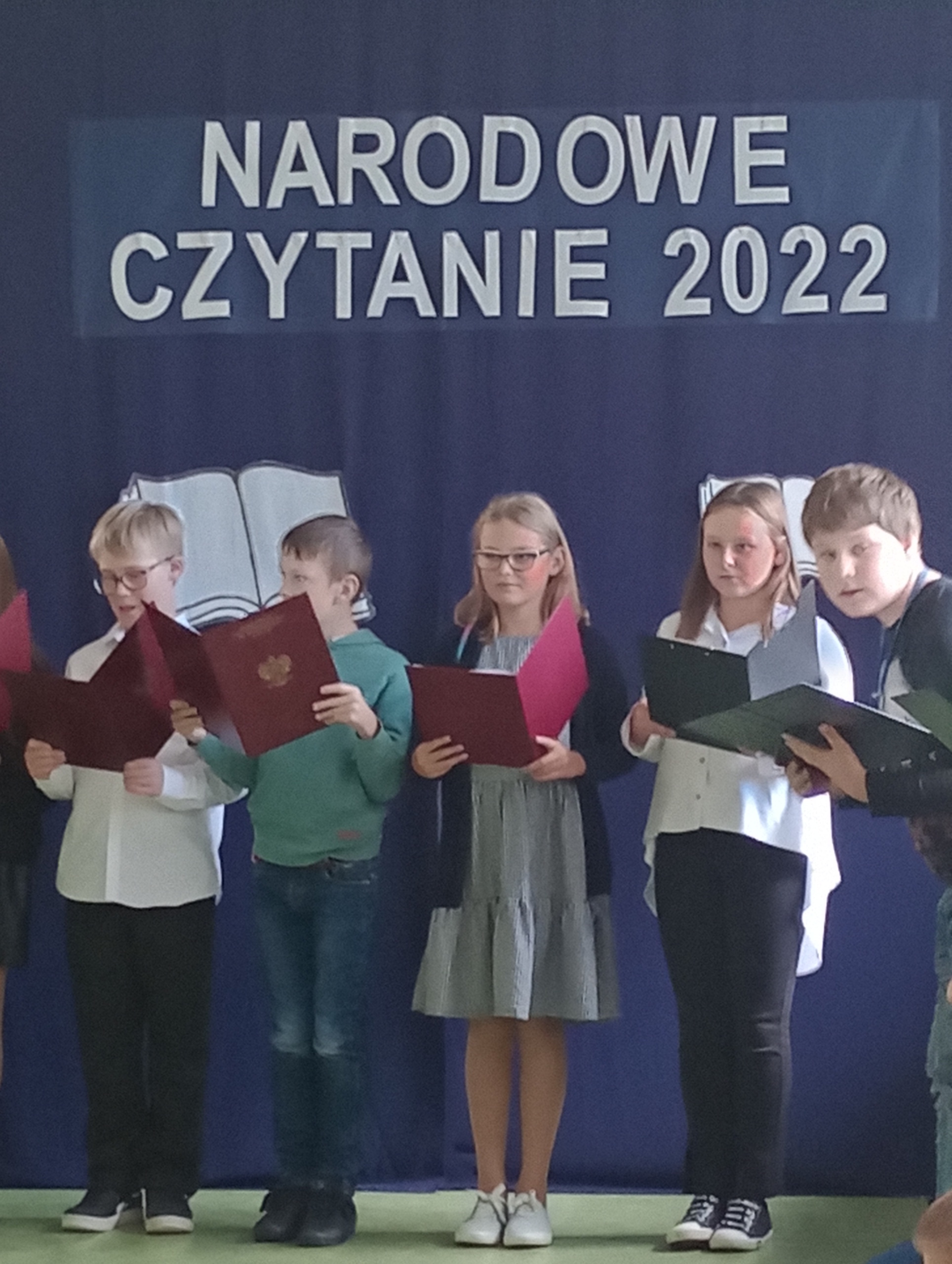 Na zdjęciu są dzieci, które czytają tekst lektury , w tle jest napis Narodowe czytanie 2022.