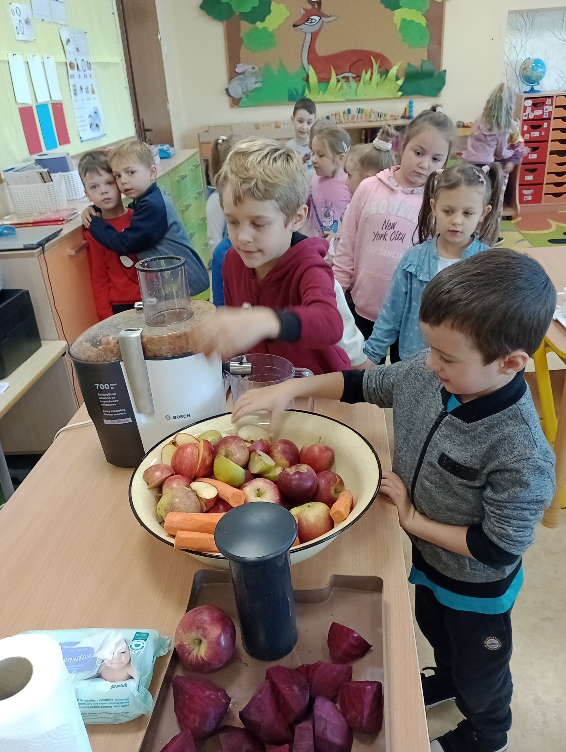 Na zdjęciu widać jak dzieci wrzucają owoce i warzywa do sokownika.
