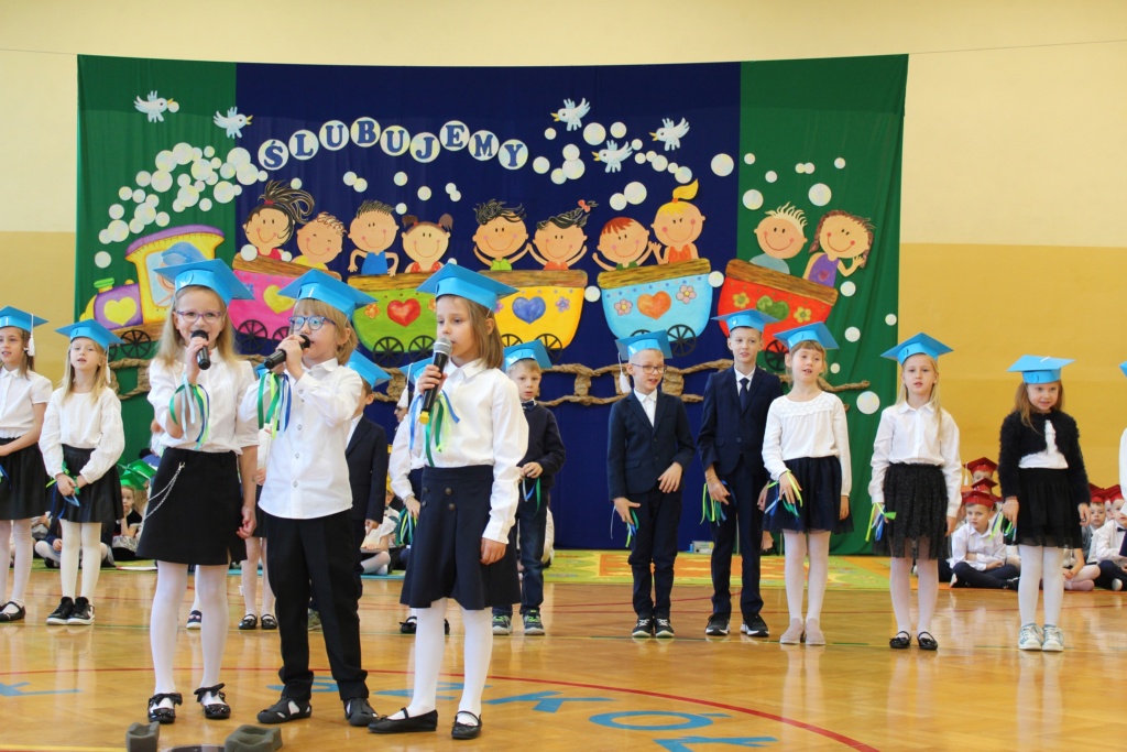 Trójka dzieci w niebieskich biretach śpiewa piosenkę ekologiczną, a reszta klasy ilustruje ją ruchem .