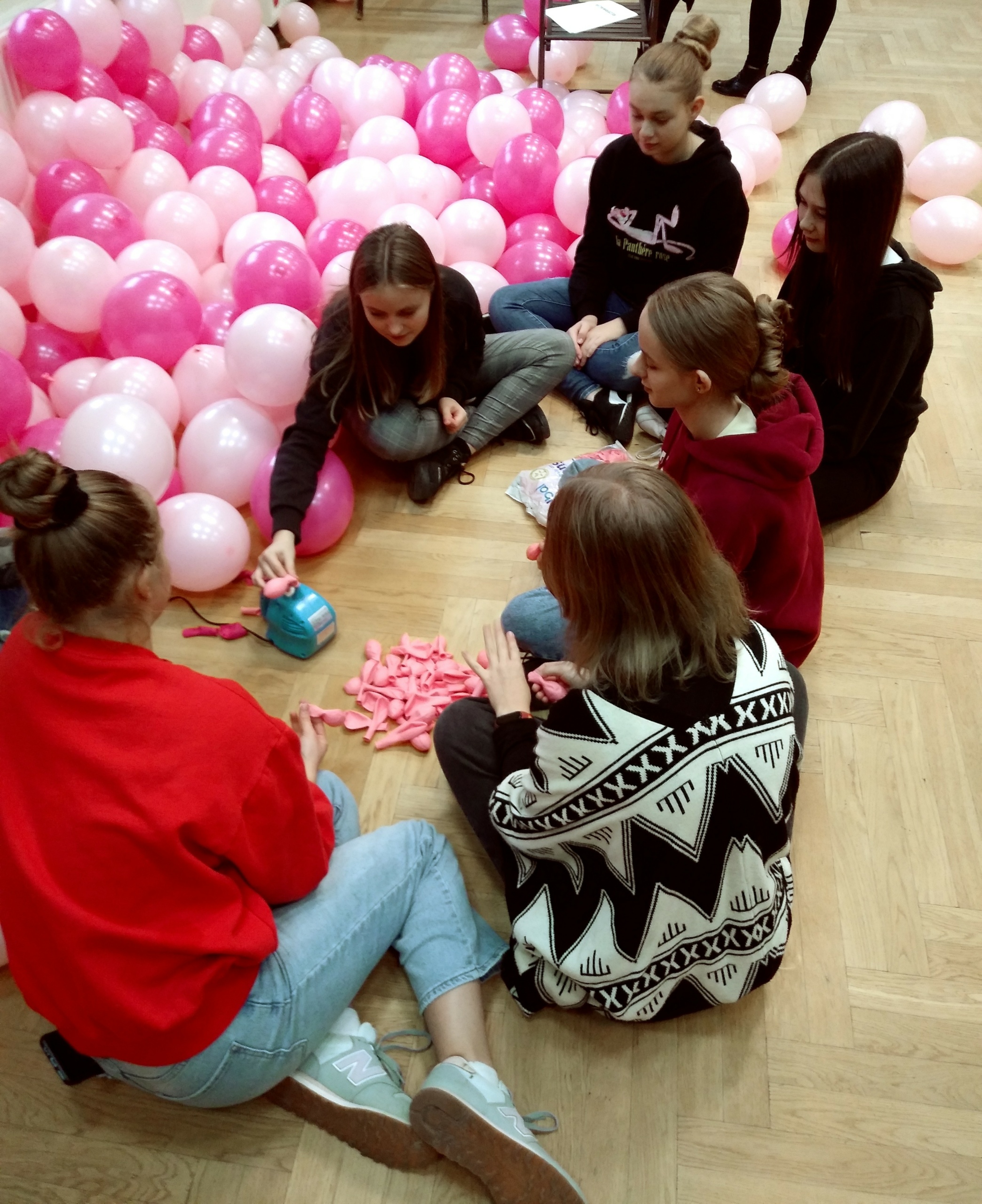 Zdjęcie przedstawia wolontariuszy, siedzących na podłodze, w tle napompowane, różowe balony.