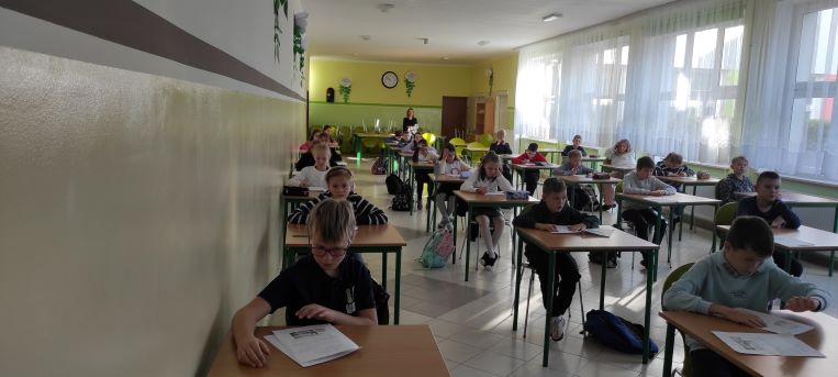 uczniowie klas IV, chłopcy i dziewczęta; pochyleni nad stolikami rozwiązują zadania testowe; miejsce - stołówka szkolna