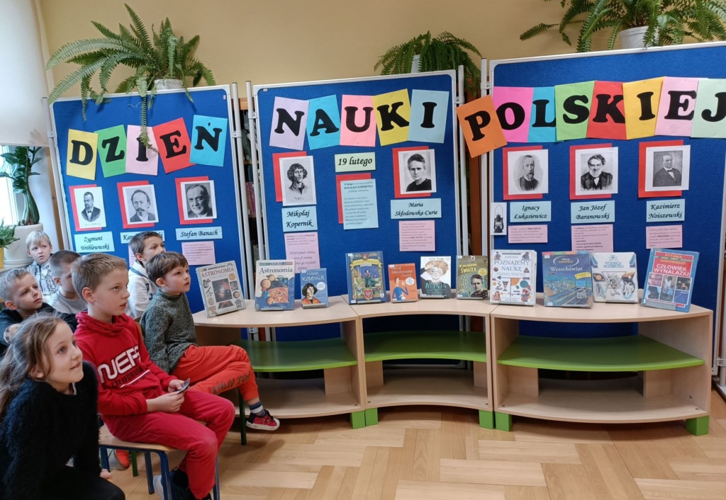 uczniowie klasy 2d na spotkaniu z okazji Dnia Nauki Polskiej. W tle widać gazetkę o słynnych naukowcach oraz wystawę książek 