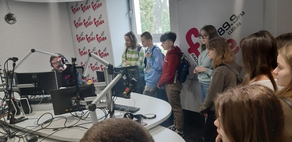 Dzieci znajdują się w studiu nagrań gdzie program prowadzi radiowiec.  Na środku widoczne jest duże białe biurko, na którym  są komputery i mikrofony, po prawej stronie stoją uczniowie.