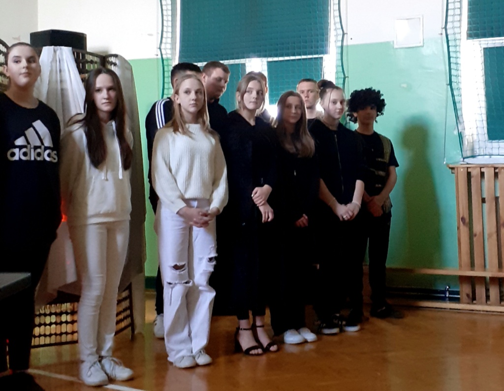 Uczniowie z grupy realizującej projekt stoją w rzędzie na sali gimnastycznej. Ubrani są na czarno lub na biało.