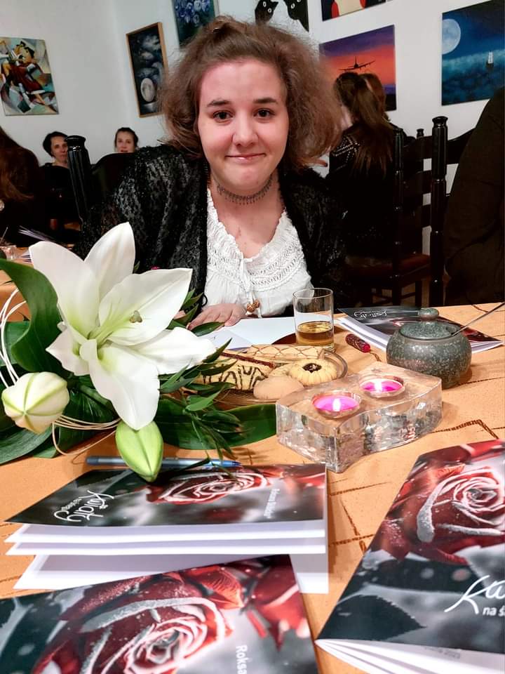 Bohaterka wieczoru- Roksana Skibicka siedzi przy stole, na który leżą lilie oraz tomiki jej poezji, pali się świeczka.