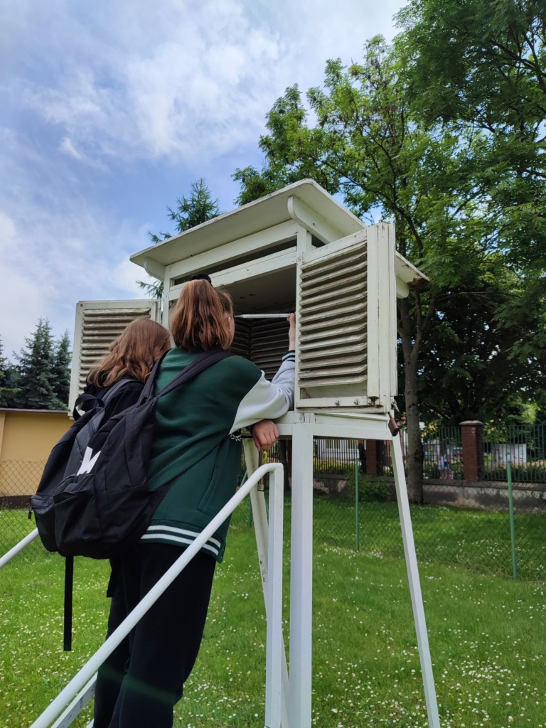 Uczennice stoją na schodkach budki meteorologicznej i sprawdzają temperaturę powietrza odczytując informację z termometru.