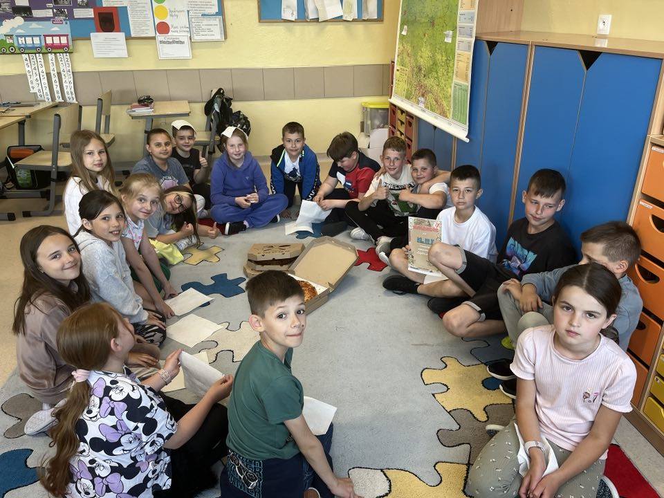 Na zdjęciu widać uczniów jednej z wyróżnionych klas w konkursie. Dzieci siedzą w kole, a  nagroda – pizza jest w środku koła. Jeden z uczniów trzyma dyplom. Wszyscy są uśmiechnięci.