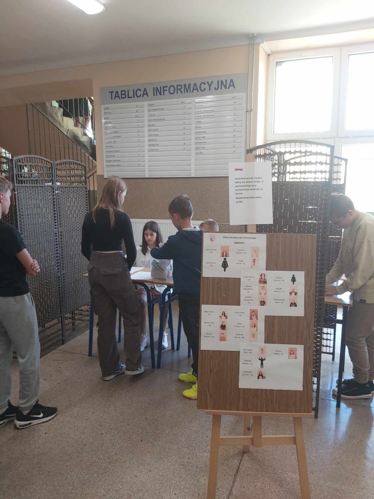 Na pierwszym planie stoi tablica ze zdjęciami kandydatów do Samorządu Uczniowskiego, w tle widać punkt wyborczy, komisja siedzi za stołem i wydaje karty do głosowania.