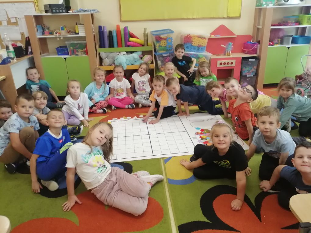 Dzieci  uśmiechnięte siedzą na kolorowym dywanie wokół maty do kodowania.