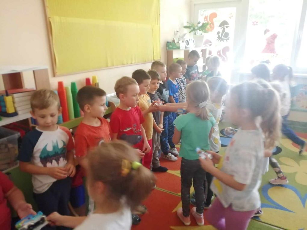 Grupa dzieci stoi w sali lekcyjnej, dziewczynki wręczają upominki chłopcom z okazji dnia chłopaka.