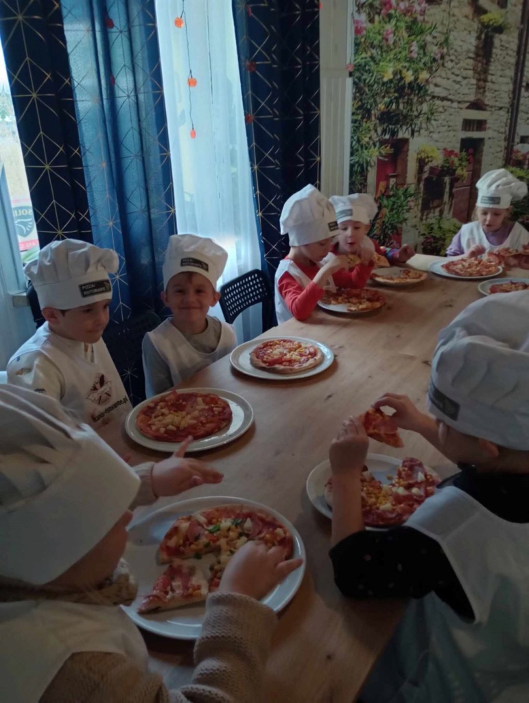 Grupa dzieci siedzi przy stolikach, na talerzach maja wykonane przez siebie pizzę. 
