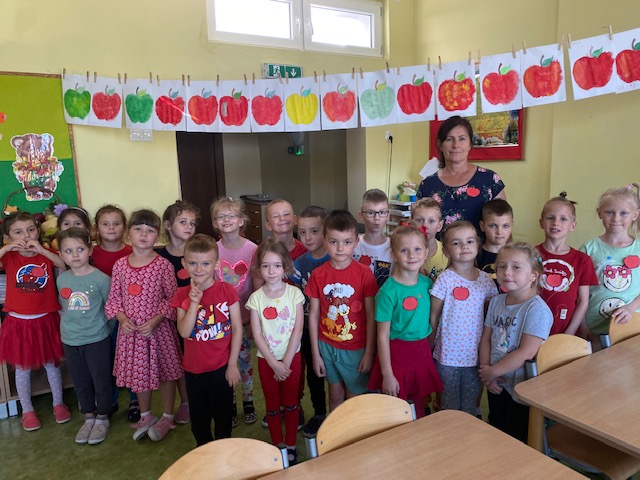 Grupa dzieci ubranych w kolory jabłka stoi z nauczycielem a nad nimi dekoracja z kolorowych jabłek.