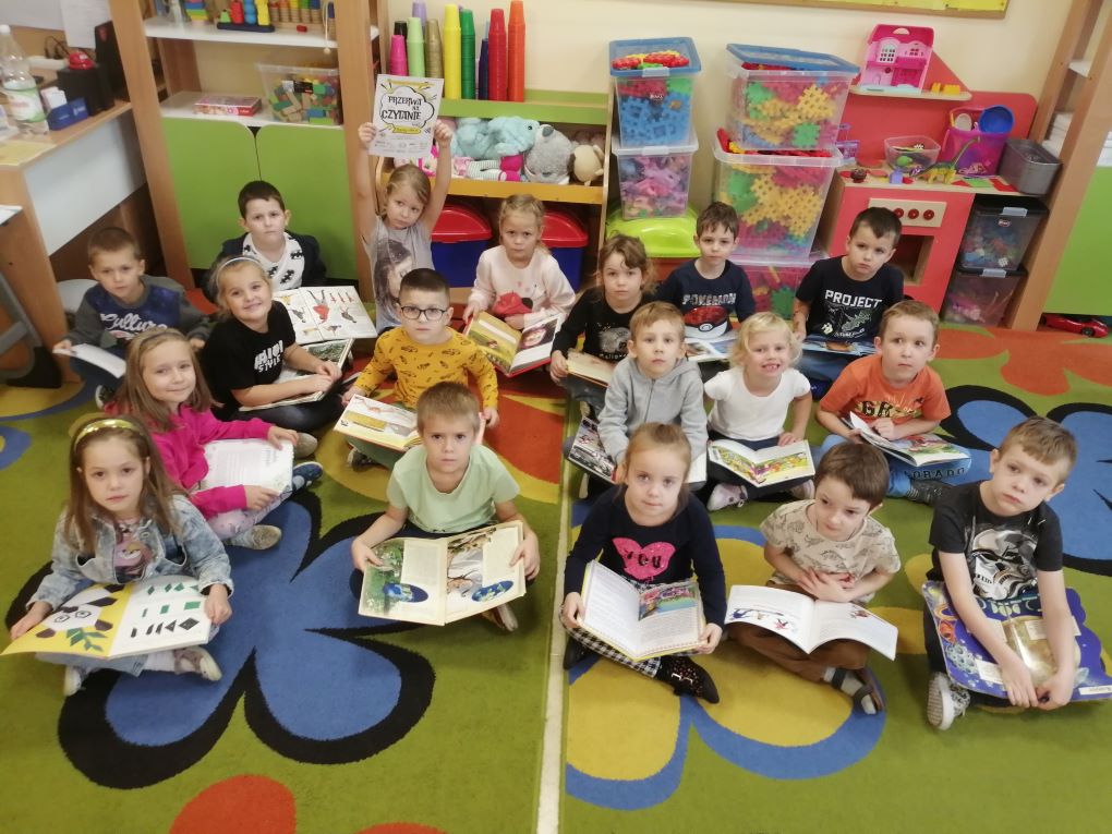 Dzieci uśmiechnięte siedzą na kolorowym dywanie i czytają książki.