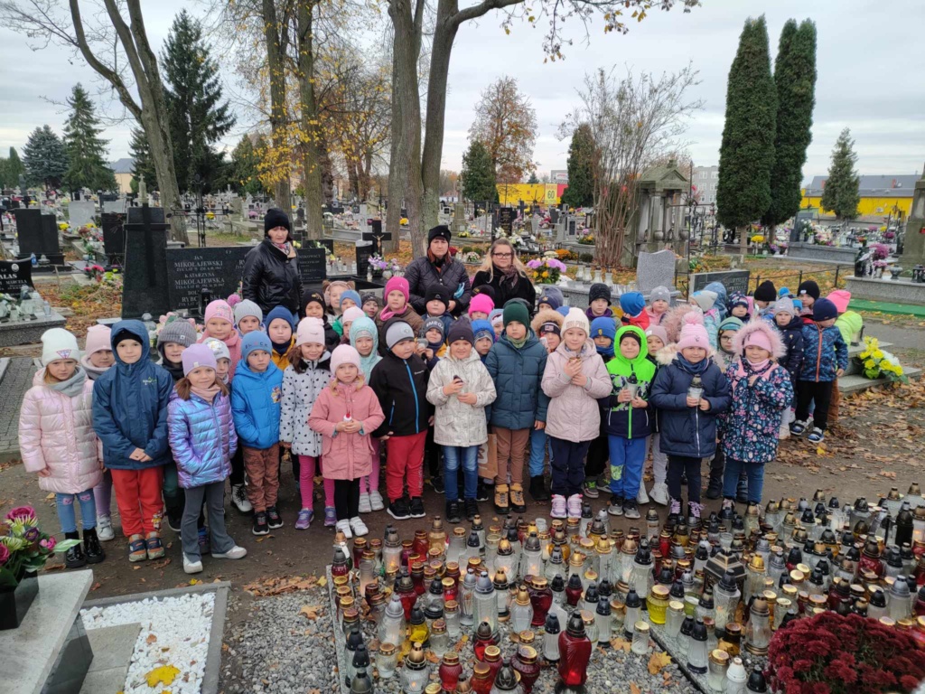 Dzieci uśmiechnięte stoją w kolorowych kurtkach na cmentarzu.