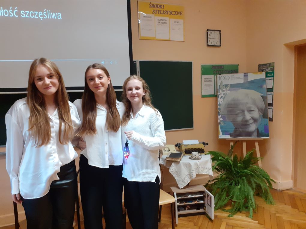 Trzy uczennice stoją przed tablicą w tle zdjęcie Wisławy Szymborskiej. 
