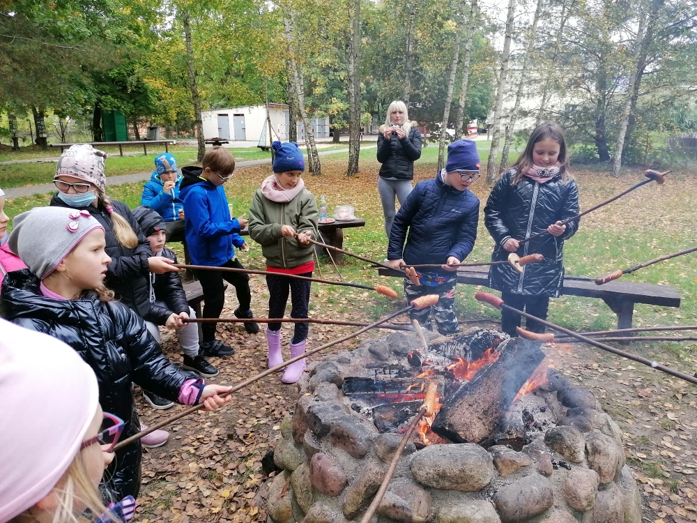 Kilkoro dzieci stoi z kijkami nad ogniskiem i piecze kiełbaski na terenie parku