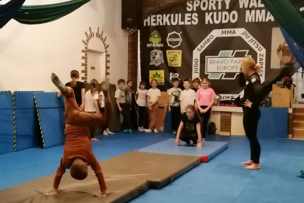 uczeń wykonuje stanie na rękach na materacu. W tle inni uczniowie przygotowują się do ćwiczenia