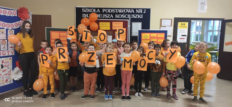 Zdjęcie 5-przedstawia klasę 1b, dzieci stoją wraz z nauczycielem, wszyscy są ubrani na pomarańczowo, w dłoniach trzymają pomarańczowe balony lub pomarańczowe kartki z literami tworzącymi hasło STOP PRZEMOCY