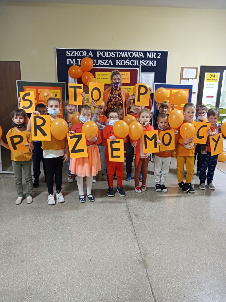 Zdjęcie 7 - przedstawia klasę 1d, dzieci stoją wraz z wychowawcą, wszyscy są ubrani na pomarańczowo, w dłoniach trzymają pomarańczowe balony lub pomarańczowe kartki z literami tworzącymi hasło STOP PRZEMOCY