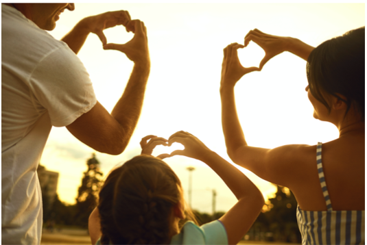 Zdjęcie przedstawiające trzyosobową rodzinę odwróconą ku słońcu gestami rąk pokazującymi kształty serc. 