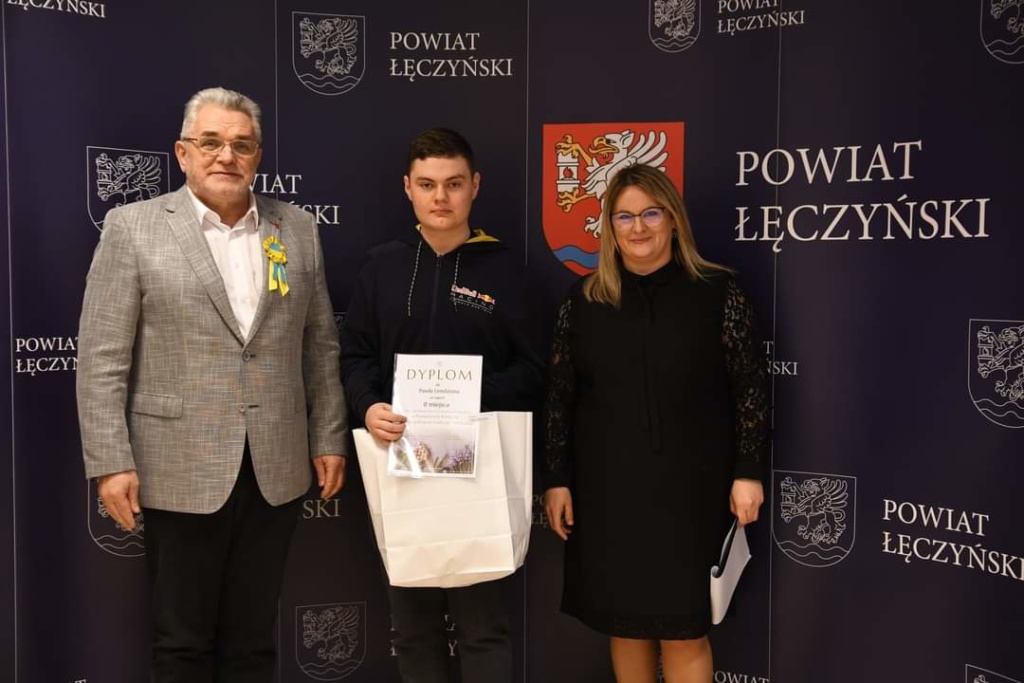 Na zdjęciu z lewej strony jest laureat konkursu Paweł Lendzion ubrany w granatową bluzę, odbierający nagrodę. W środku jest pan Starosta łęczyński ubrany w szarą marynarkę, a z prawej strony pani naczelnik - jedna z głównych organizatorów konkursu.