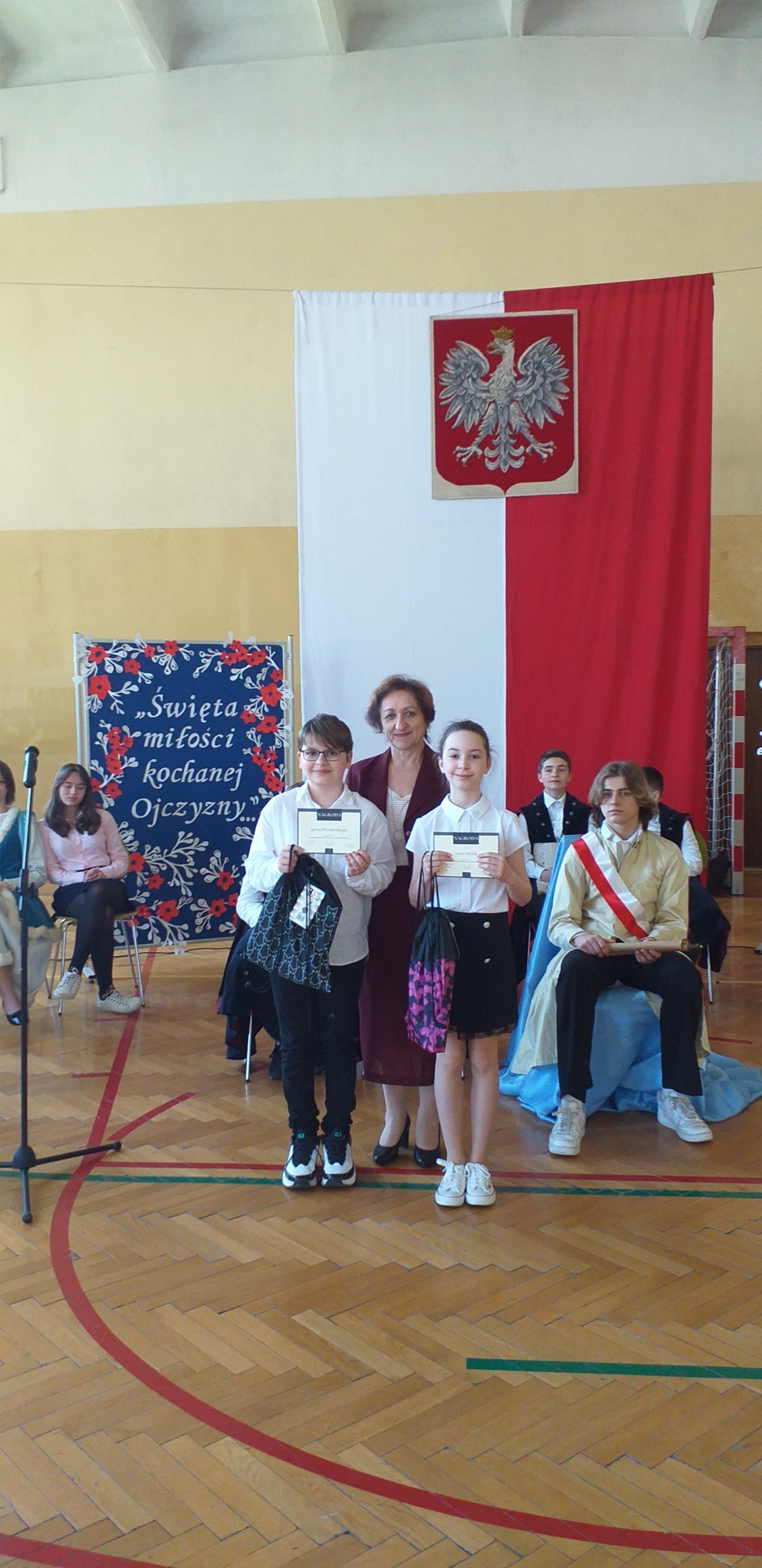 Czarne spodnie, obok niego stoi kobieta w czarnym żakiecie i dziewczyna w białej bluzce i granatowej spódnicy, dzieci w rękach trzymają dyplomy.