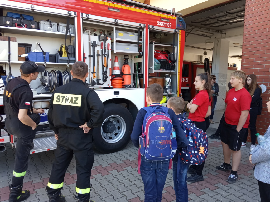 Dwóch strażaków opowiada i pokazuje uczniom samochód i sprzęt strażacki.