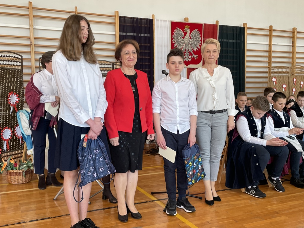 Grupa, w pierwszym planie od lewej stoi wysoka dziewczyna ubrana w białą bluzkę i granatową spódnicę, ma długie włosy, obok niej stoi kobieta ubrana w czarną sukienkę i na niej ma czerwony żakiet, chłopiec w białej koszuli i w czarnych spodniach i kobieta w białej bluzce i spodniach w biało, niebieskie prążki.
