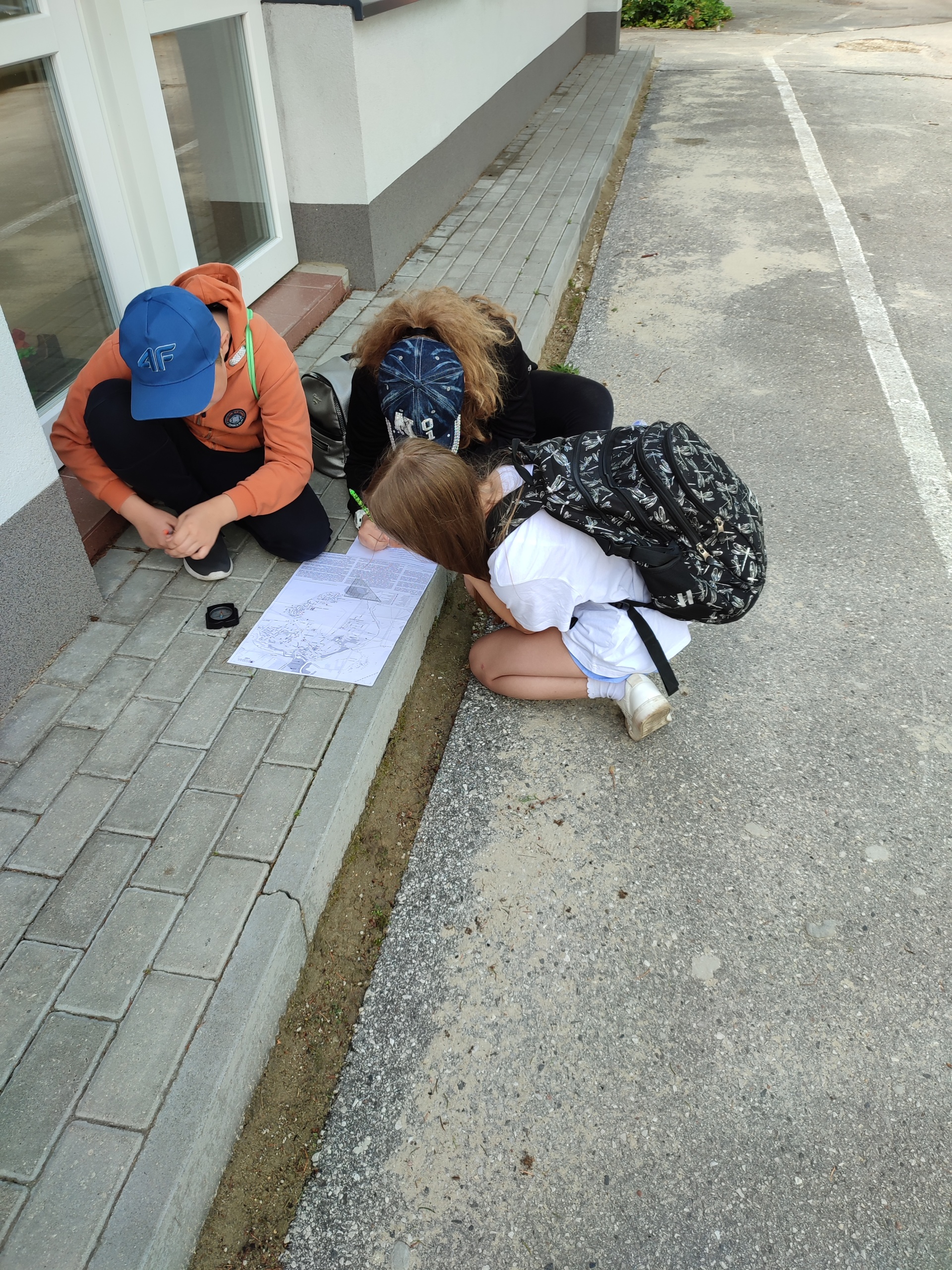 Trzech uczniów w róznokolorowych strojach z plecakami na chodniku, obok białego budynku, ogląda na klęcząco mapę. Obok leży kompas. 