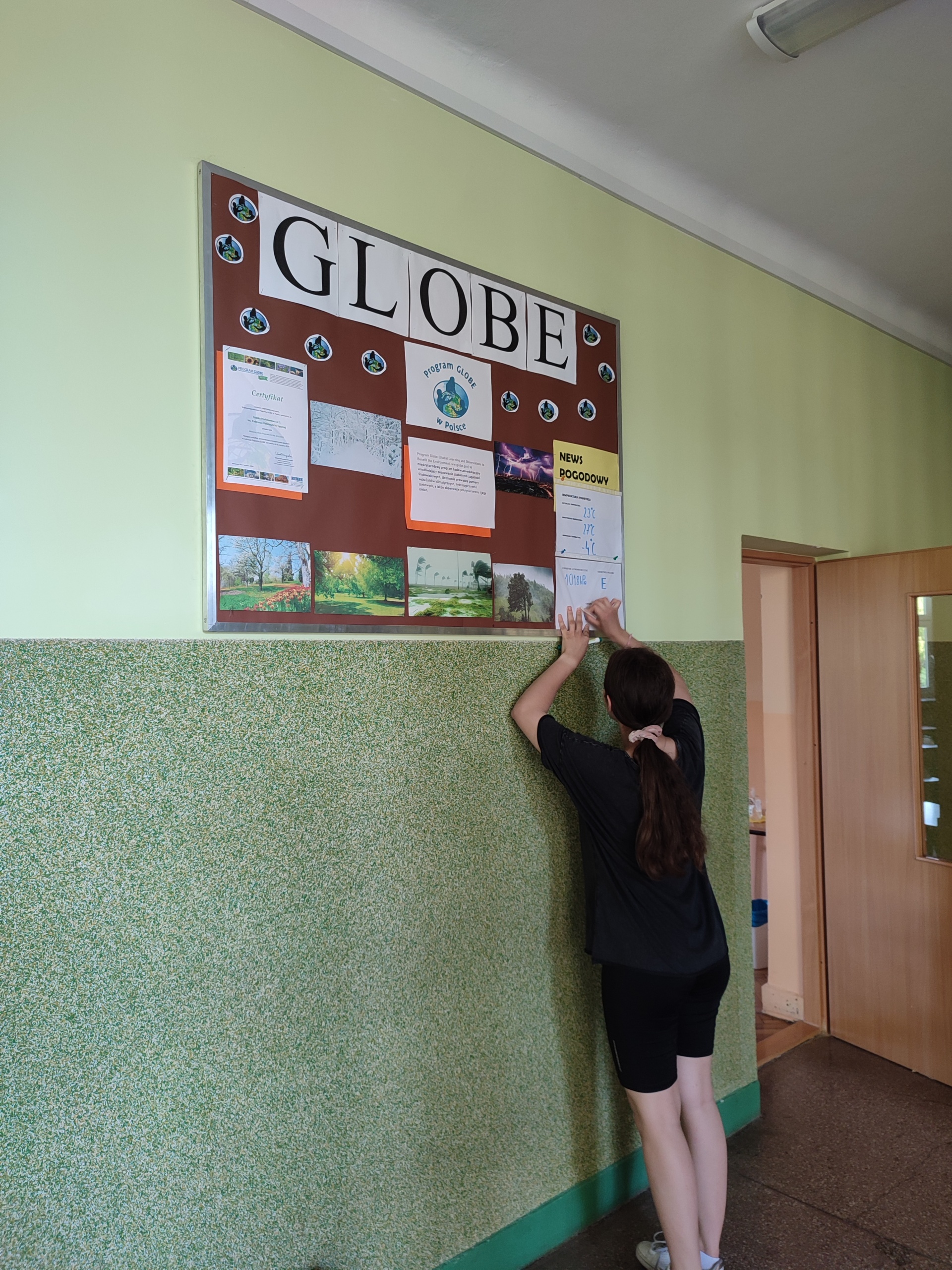Wnętrze szkoły, korytarz, na zielonej ścianie wisi tablica z napisem GLOBE, na niej róznokolorowe plansze. Uczennica ubrana na czarno przyczepia na tablicy nową planszę. W oddali widać fragment brązowych drzwi. 