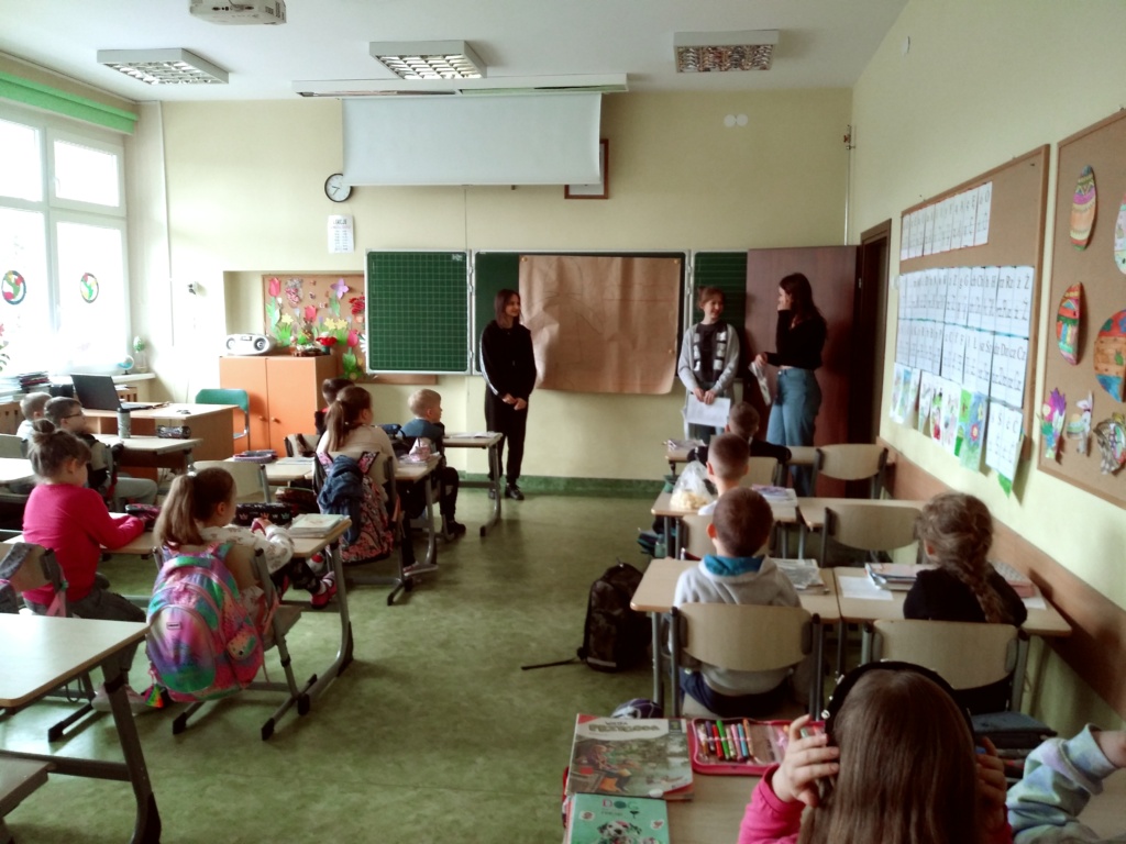 Zdjęcie przedstawia uczniów w sali lekcyjnej, wolontariuszki przy tablicy, młodszych uczniów w ławkach.