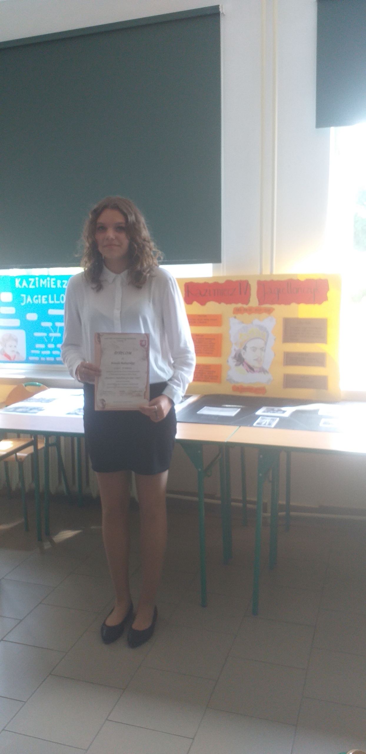 Na zdjęciu dziewczyna z długimi, kręconymi włosami, za nią żółty plakat z głową króla