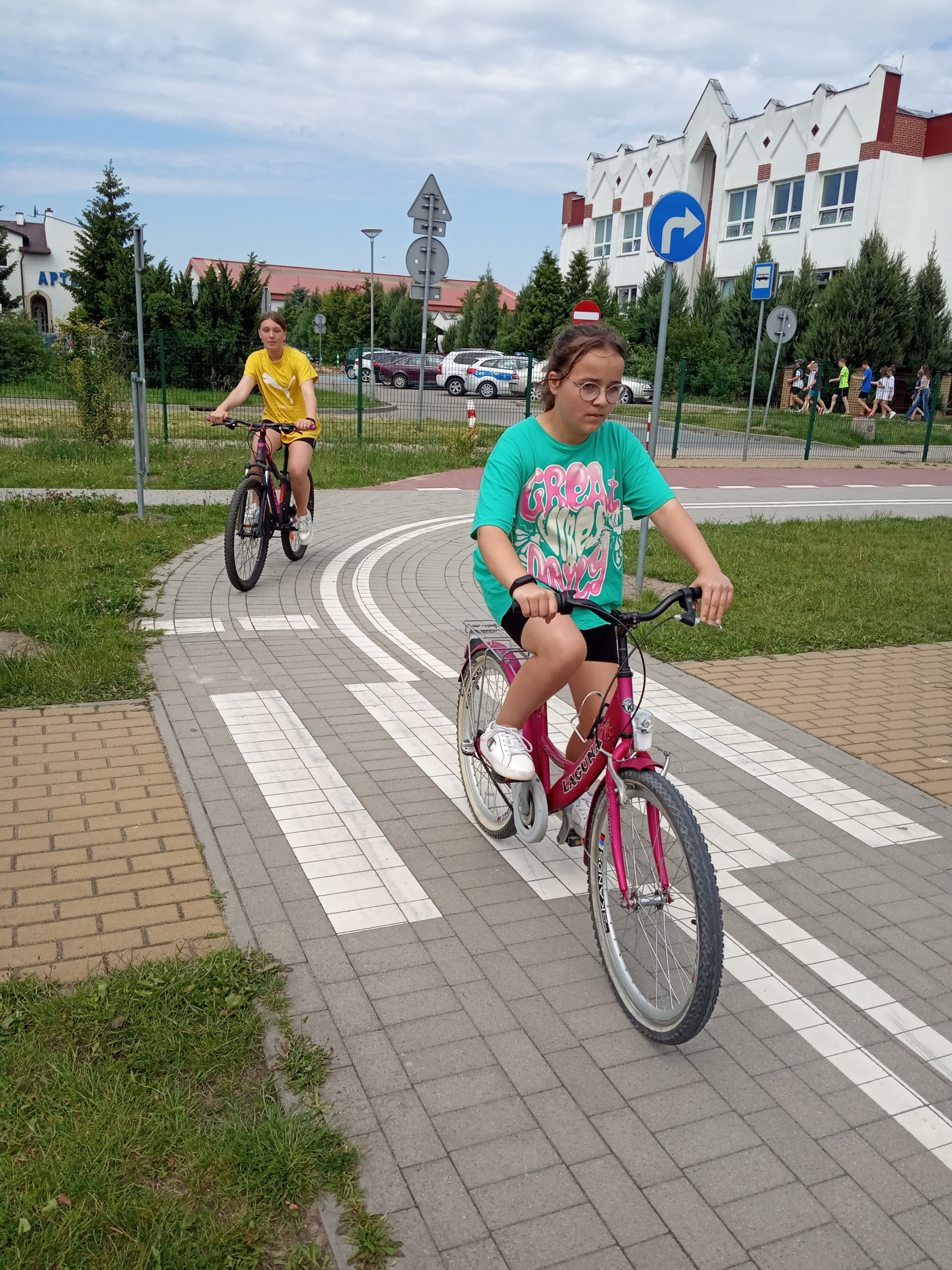 Uczennica jedzie rowerem przez skrzyżowanie, za nią jedzie kolejna osoba.