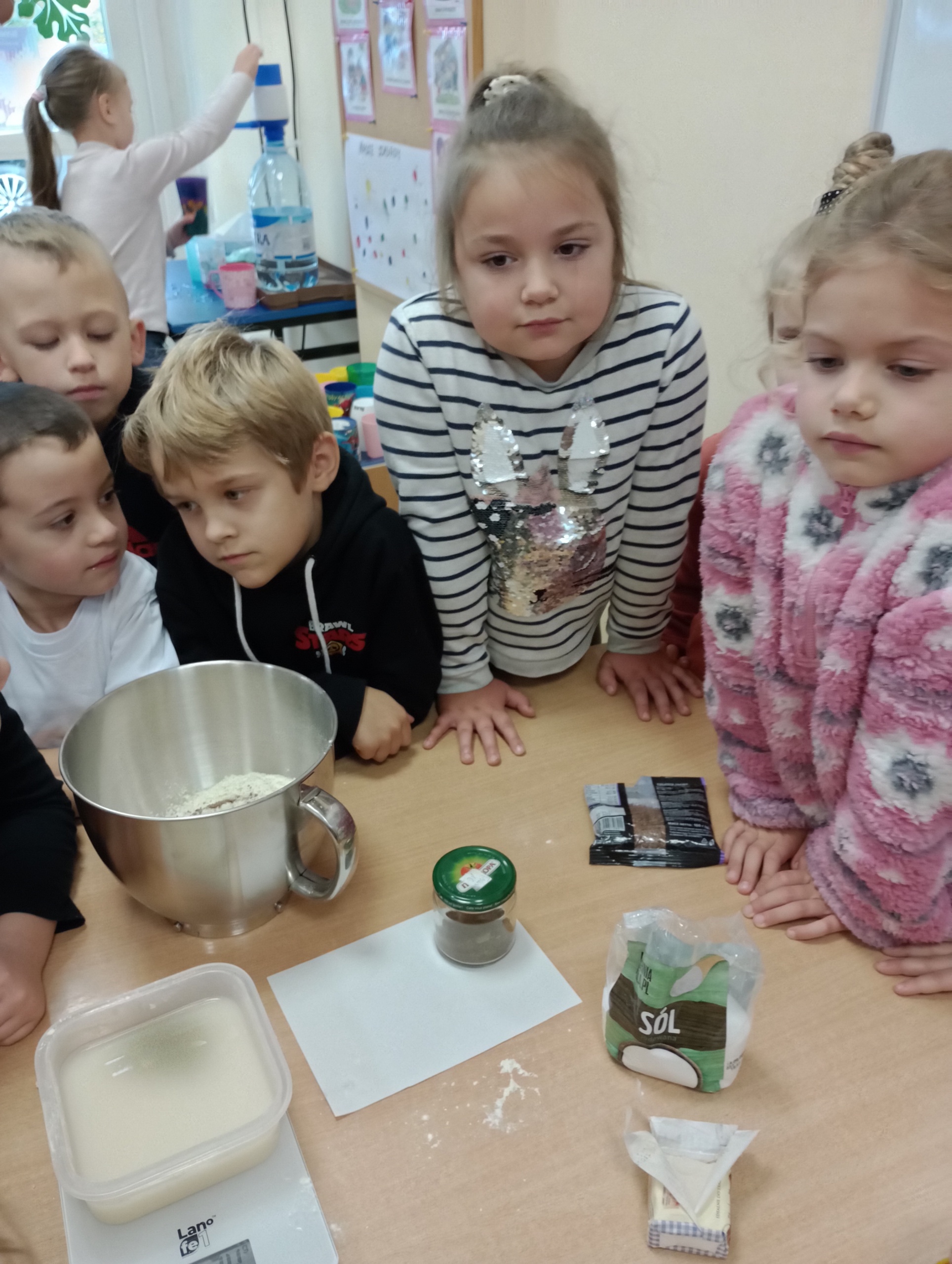 dzieci stoją przy stoliku, na którym znajdują się produkty potrzebne do wypieku chleba