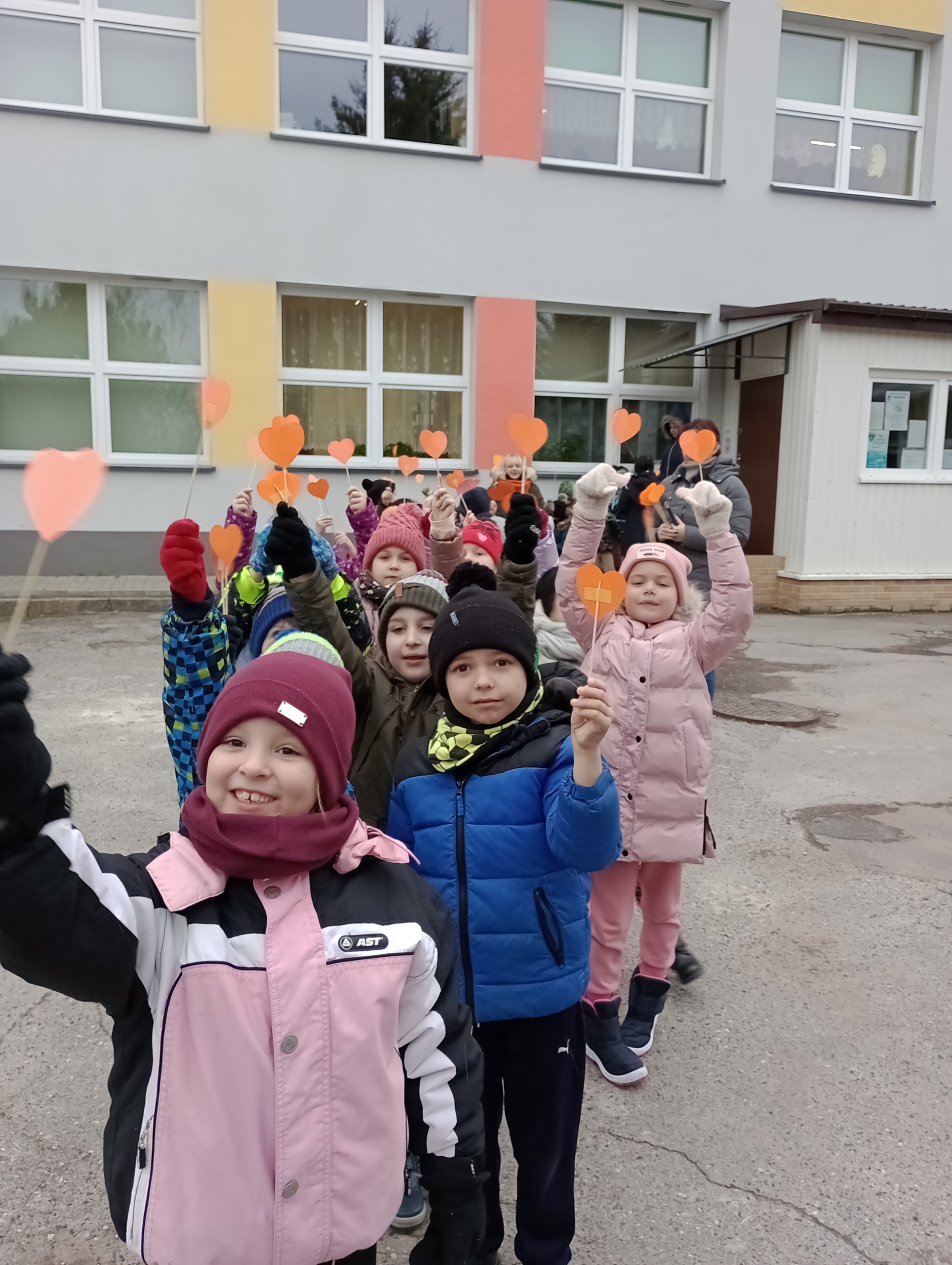 Na zdjęci dzieci stoją w parach na dworze a w ręku trzymają pomarańczowe serduszka