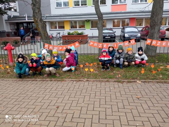zdjęcie przedstawia grupę przedszkolną przed budynkiem szkoły przy transparencie Kampania Przeciwko Przemocy