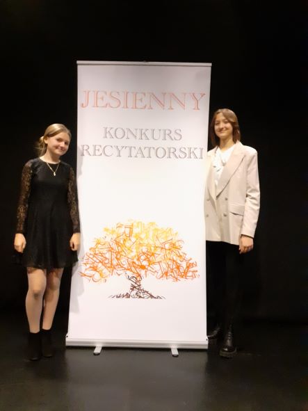 na zdjęciu laureatki konkursu, uczennice Emilia Cegłowska i Milena Sobiesiak; uczennice są uśmiechnięte; stoją przy banerze Jesiennego Konkursu Recytatorskiego;