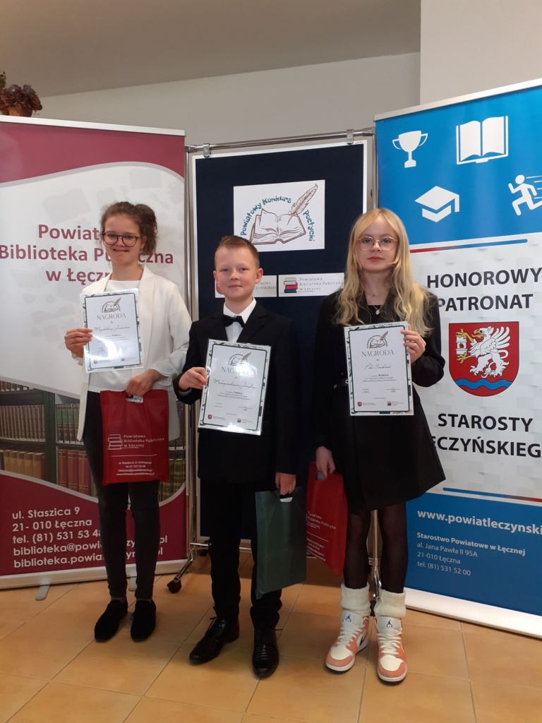  laureaci konkursu stoją przed banerami Powiatowej biblioteki Publicznej w Łęcznej i Powiatu Łęczyńskiego, w rękach trzymają pamiątkowe dyplomy i nagrody.