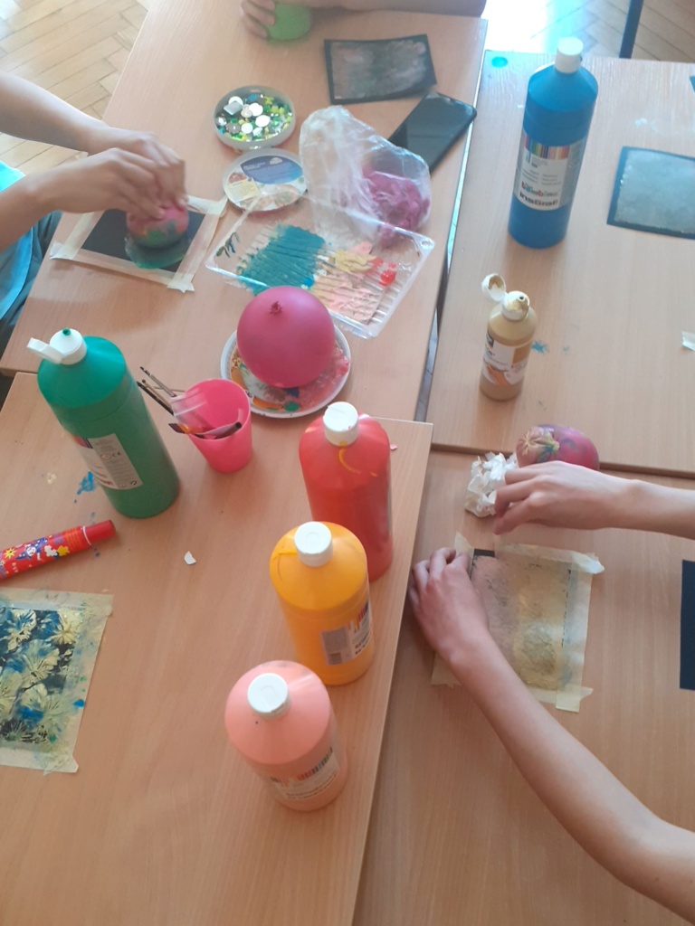 uczniowie siedzą przy stole i wykonują grafiki przy pomocy balonu moczonego w farbach akrylowych