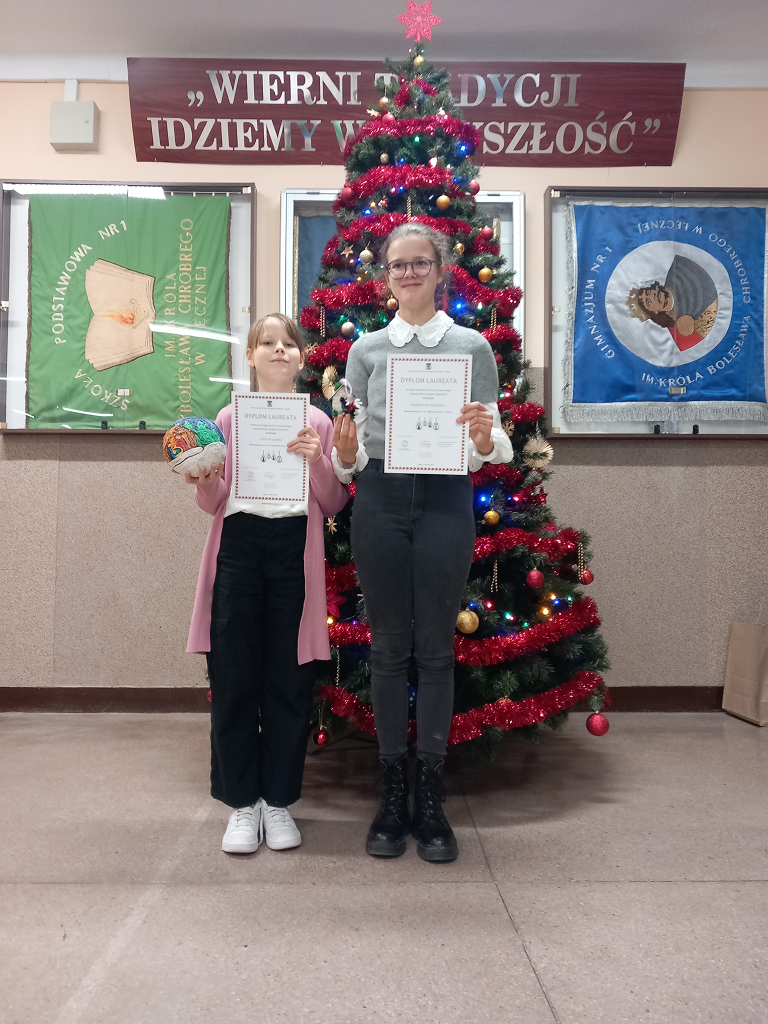 Na tle choinki stoją Magdalena Jakubiec oraz Julia Piasecka laureatki Wojewódzkiego Konkursu Świątecznego „Najpiękniejsza bombka choinkowa”. W rękach trzymają dyplomy i nagrodzone prace.