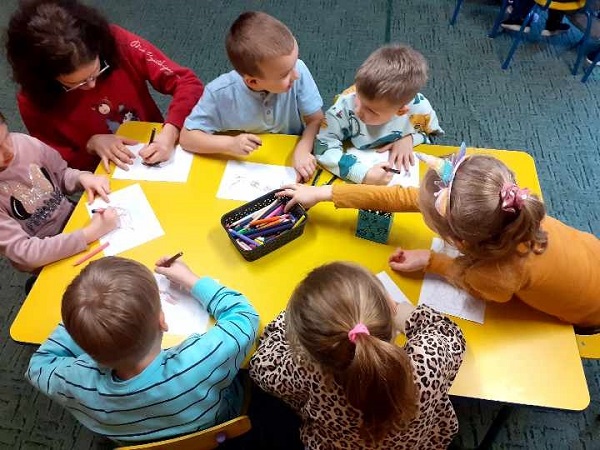 Dzieci siedzą przy stoliku i kolorują obrazki. W tym samym czasie rozmawiają ze sobą.