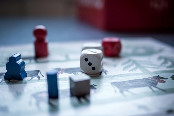 Zdjęcie przedstawia kostkę i pionki na tle gry planszowej.