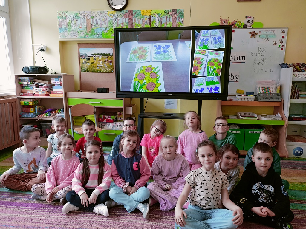 Uśmiechnięte dzieci siedzą na dywanie przed monitorem interaktywnym, na którym znajduje się wiosenny kolaż.